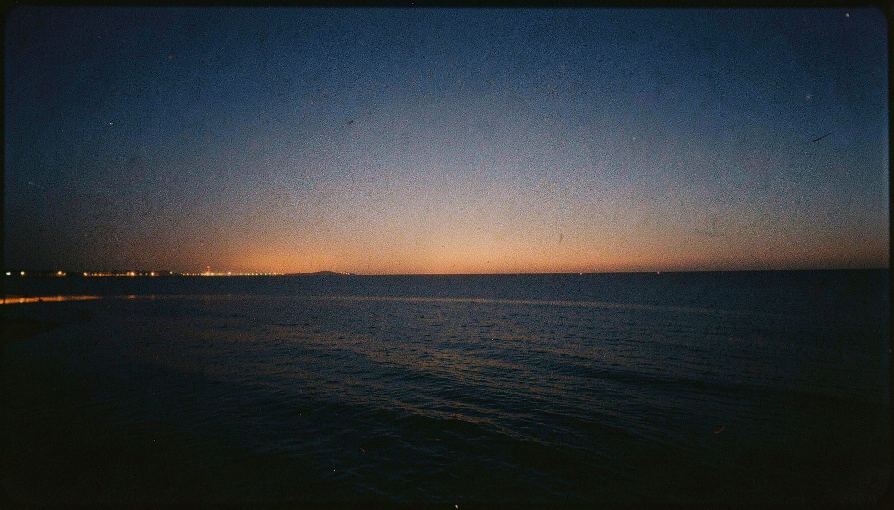 A wide analog film photograph of the Arabian Gulf at dusk, with warm amber light along the water and a deep navy sky, subtle film grain visible throughout.